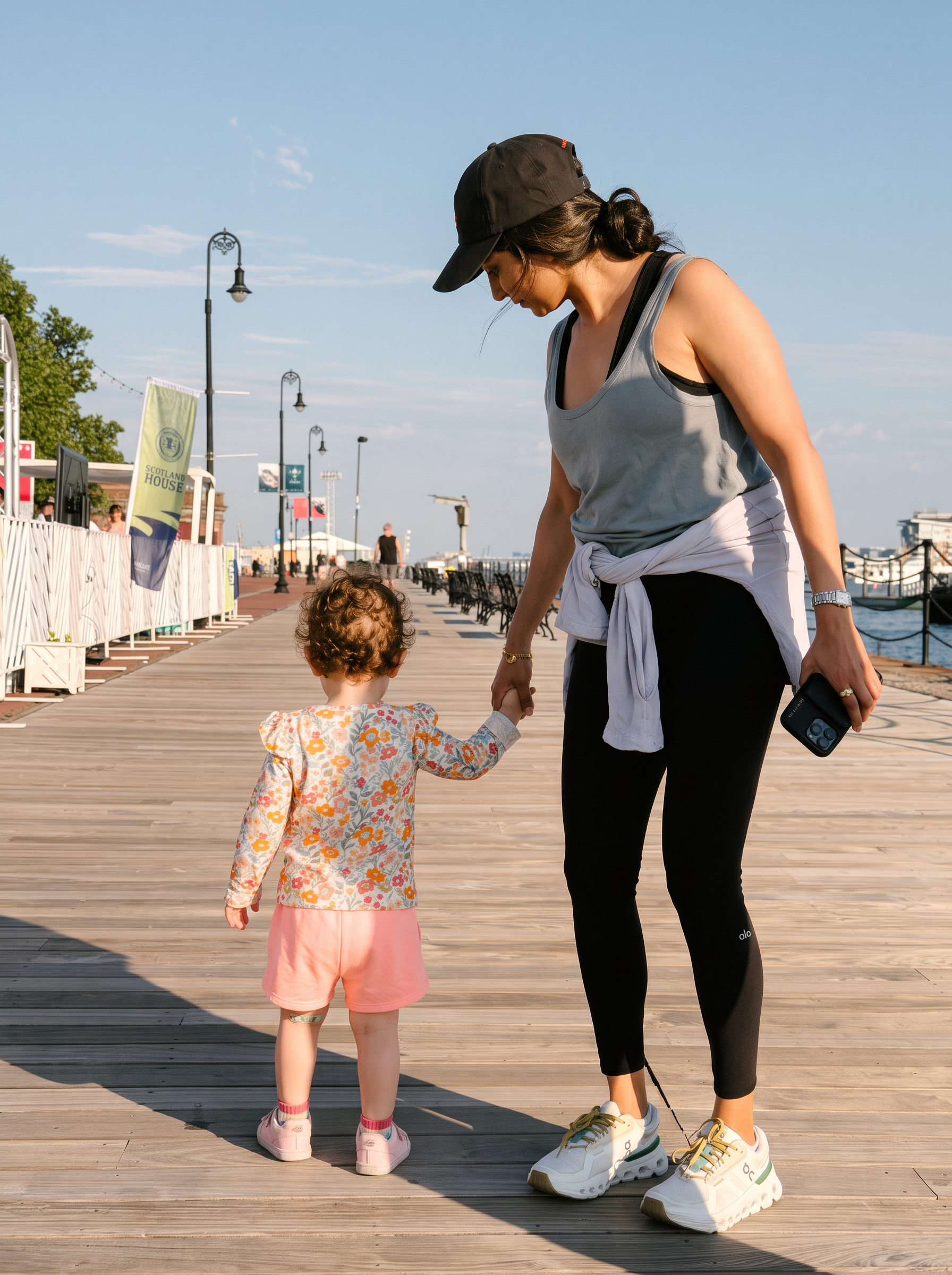 A mother walking hand in hand with her toddler along a sunlit waterfront boardwalk